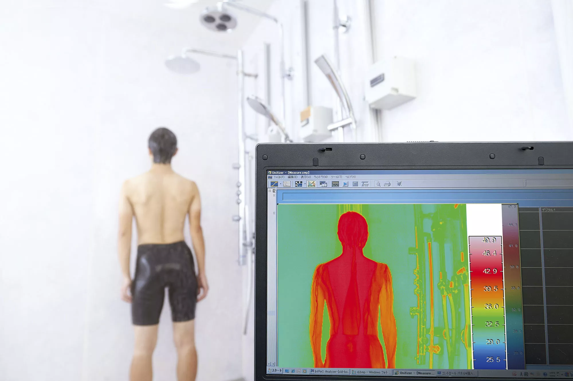 A person stands in a test shower while a nearby computer screen displays a thermal scan of their upper body heat distribution.