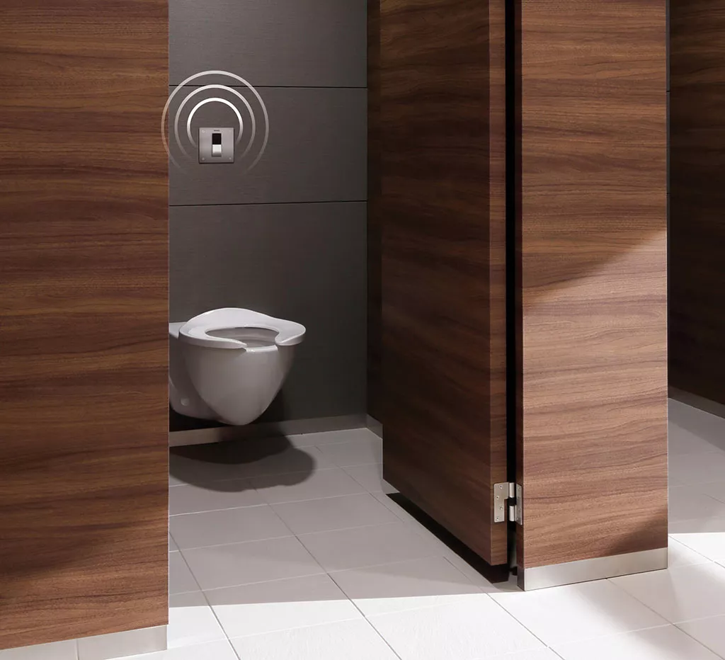 A public restroom stall with a TOTO wall-mounted toilet and IoT-enabled sensor-operated flush valve, framed by wood-finish partitions.