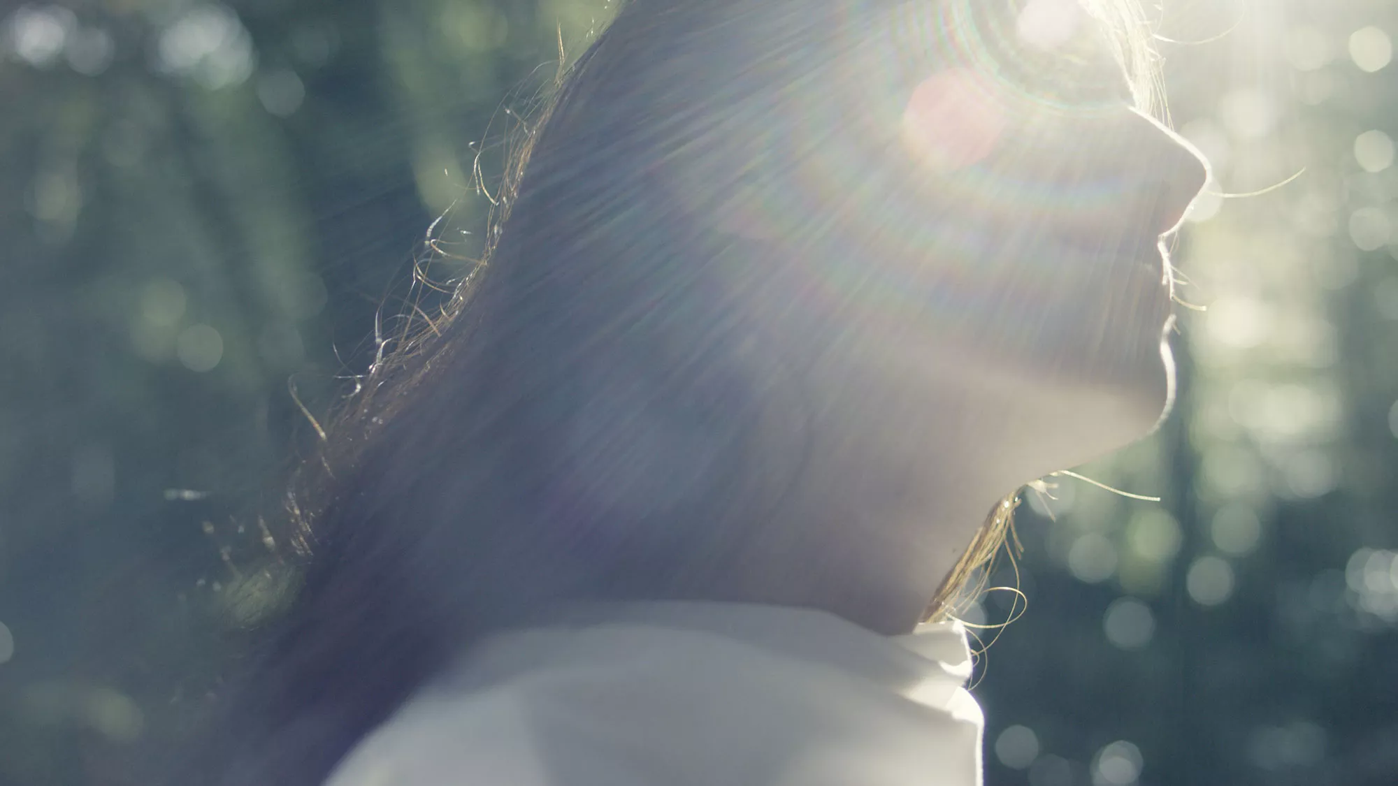 A woman tilts her face upward as soft sunlight streams through trees, casting natural light flares across her profile.